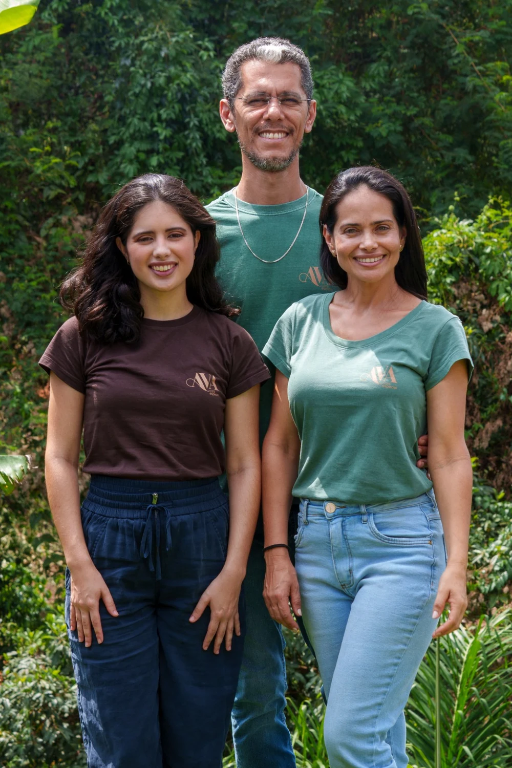 Maria Clara, Cassiano e Adélia, de camisetas da AVA, sorrindo no gramado do sítio com a mata ao fundo
